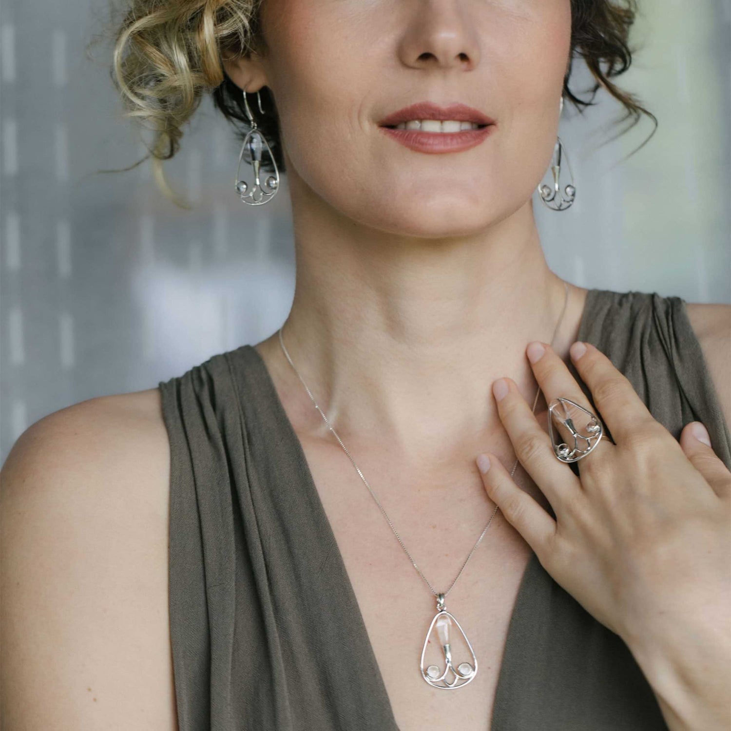 A person adorned in sophisticated jewelry from Kaora Sandara Jewelry, featuring teardrop-shaped earrings, a necklace, and a ring with Crystal Stone accents—all crafted from Sterling Silver 925—dressed in a sleeveless olive green top. Their hand gracefully touches the Self-Worth Pendant from the same collection.