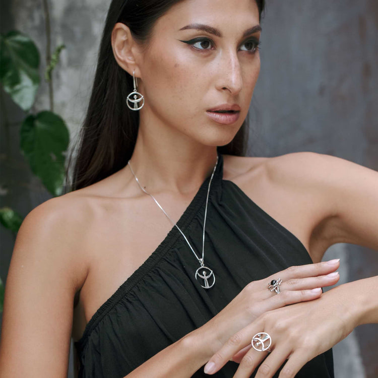 A person with long hair is wearing a black one-shoulder top, accentuated by the "Releasing From the Deepest Fears Ring" in sterling silver, size S, from Kaora Sandara Jewelry. They also showcase a matching necklace and earrings along with additional rings while posing with their left hand on their right arm against a backdrop of blurred green leaves.