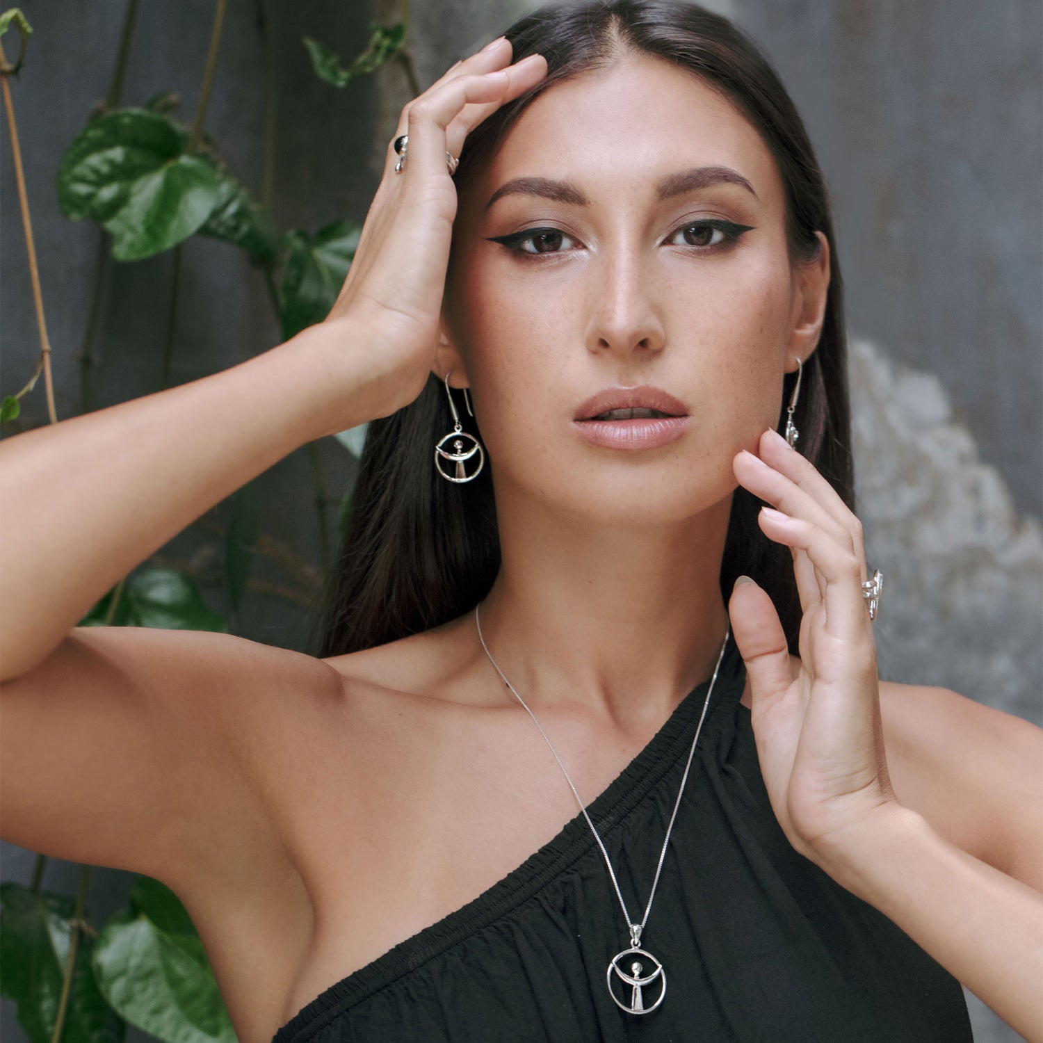 A woman with long hair poses outdoors, wearing a one-shoulder black top and accessorizing with Kaora Sandara Jewelry's "Releasing From the Deepest Fears Pendant" in sterling silver. She also wears sterling silver earrings, while green leaves and a textured wall form the background.