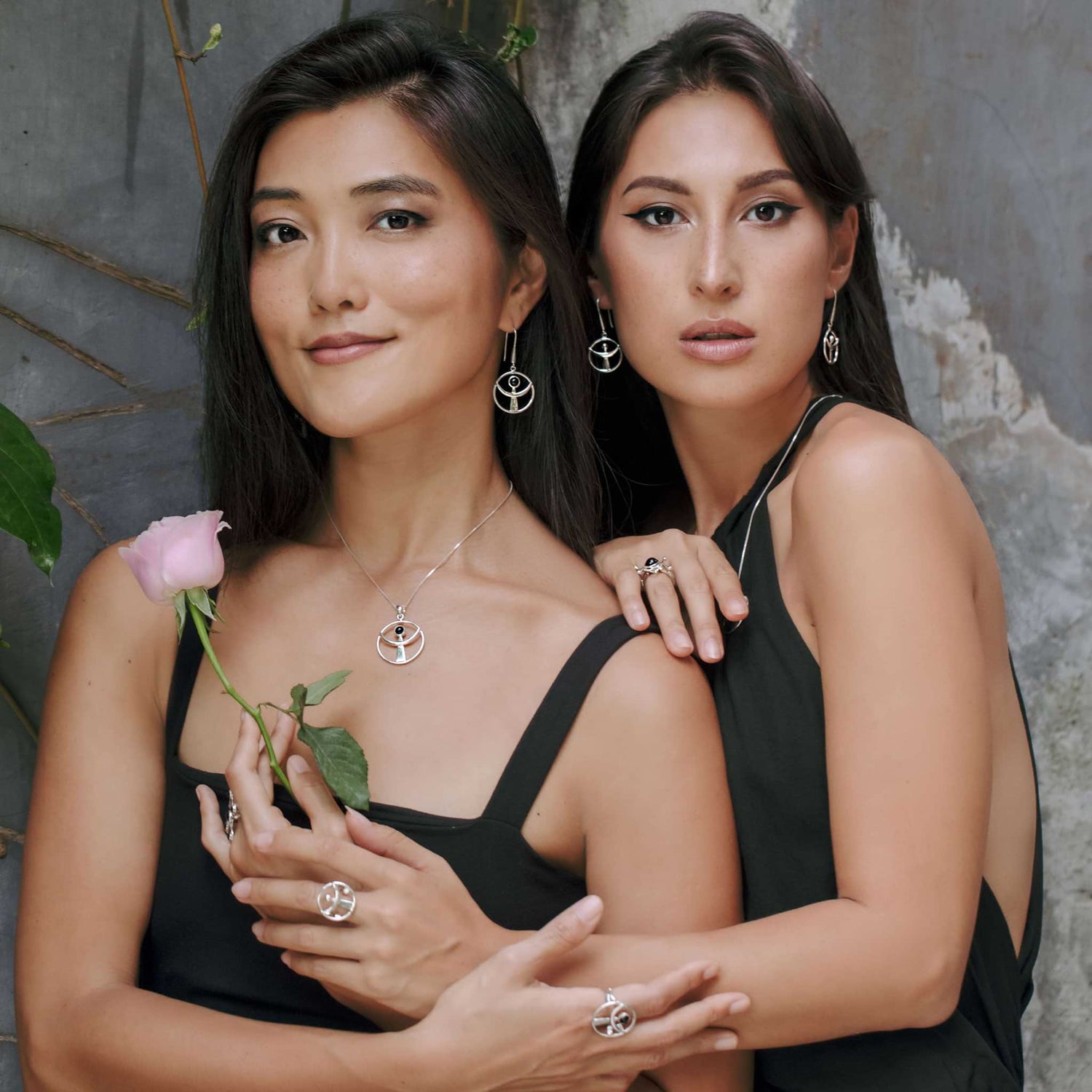 Two women in black dresses pose intimately in front of a textured wall, with one holding a pink rose. Both showcase matching jewelry, including Kaora Sandara's Sterling Silver 925 necklaces and rings featuring circular designs. Verdant leaves are visible on the left.