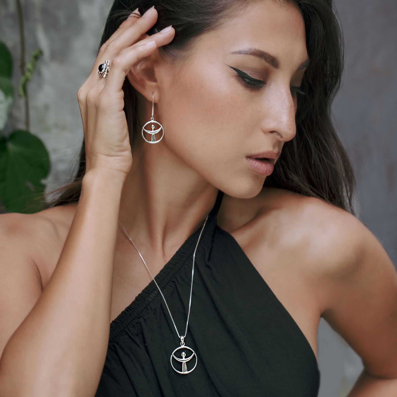 A woman with long brown hair wears a black one-shoulder dress and the Releasing From the Deepest Fears Earhooks in sterling silver by Kaora Sandara Jewelry, along with a matching necklace. She has a ring on her left hand and leans her head slightly to the side. A green plant is visible in the blurred background.