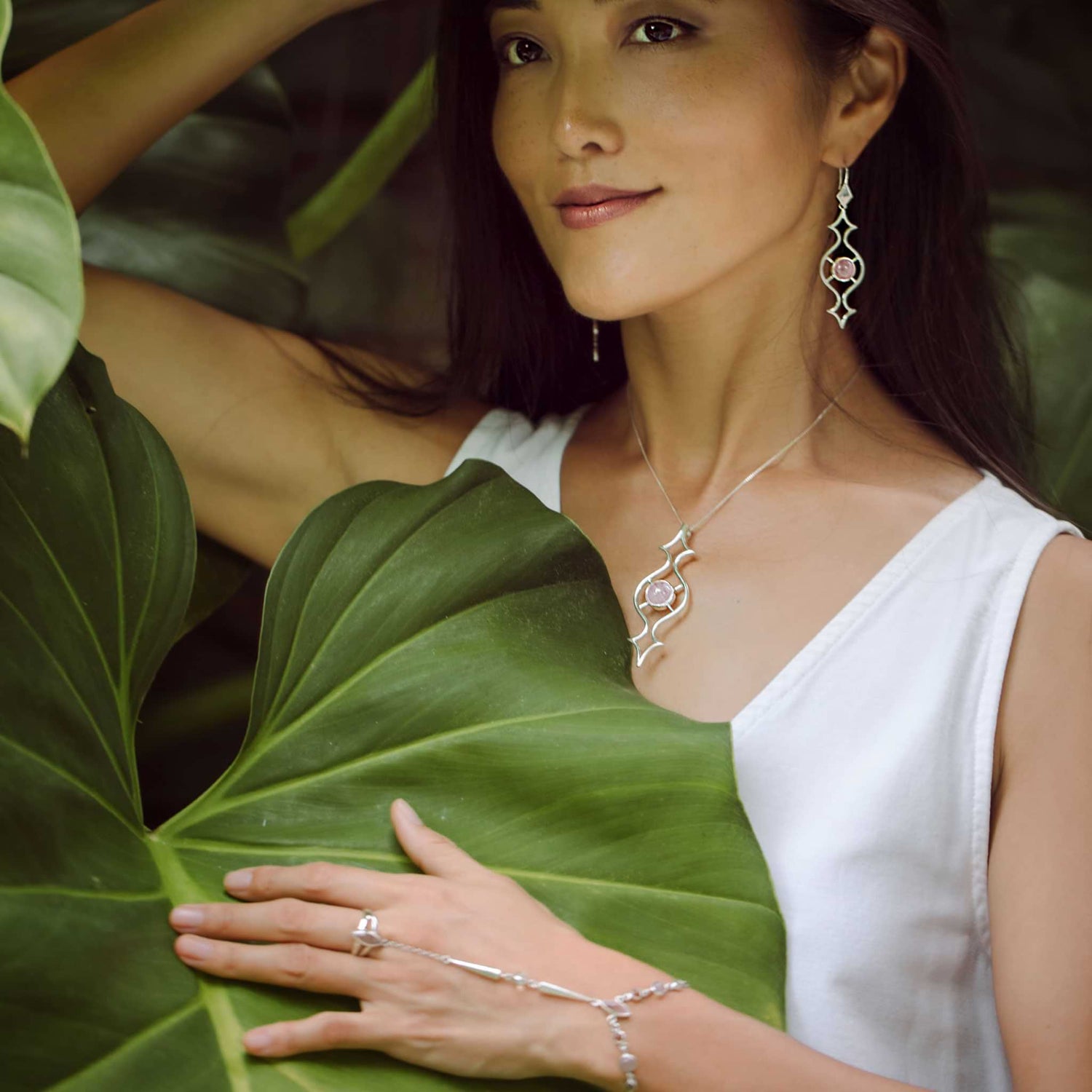 Amidst lush green leaves, a woman in a white dress elegantly displays her collection of Kaora Sandara Jewelry, featuring the Sterling Silver Quantum Shift NEW Ring with Rose Quartz in Size L. She smiles softly as her hand rests on a leaf, emphasizing the sophistication of her earrings, necklace, bracelet, and rings.