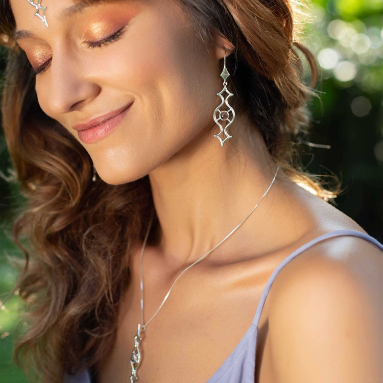 A woman with wavy brown hair smiles gently with her eyes closed, adorned in Kaora Sandara Jewelry's Quantum Shift NEW – Sterling Silver with Crystal Gemstones earrings and a matching necklace. Her warm-toned makeup complements the serene outdoor greenery as soft sunlight beautifully illuminates her face.