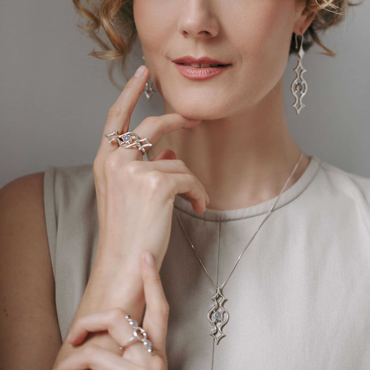 A woman in a light sleeveless top elegantly poses, showcasing intricate Sterling Silver 925 jewelry crafted by Kaora Sandara Jewelry. She's adorned with matching earrings and a necklace, along with the Quantum Shift NEW Ring with Chalcedony in size S, her hand gently touching her chin to highlight these exquisite accessories.