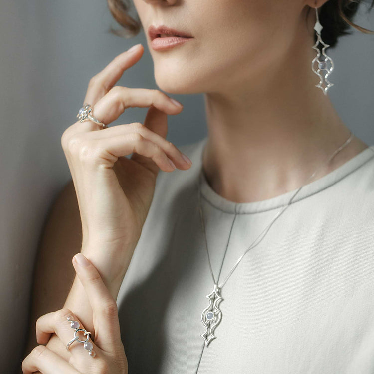 A person elegantly posed with their fingers gently touching their face, wearing an ensemble of Kaora Sandara Jewelry pieces. Adorned in the Quantum Shift NEW Earhooks crafted from Sterling Silver with Chalcedony, they are dressed in a sleeveless light gray top complemented by chalcedony accents, showcasing a subtle yet refined elegance.