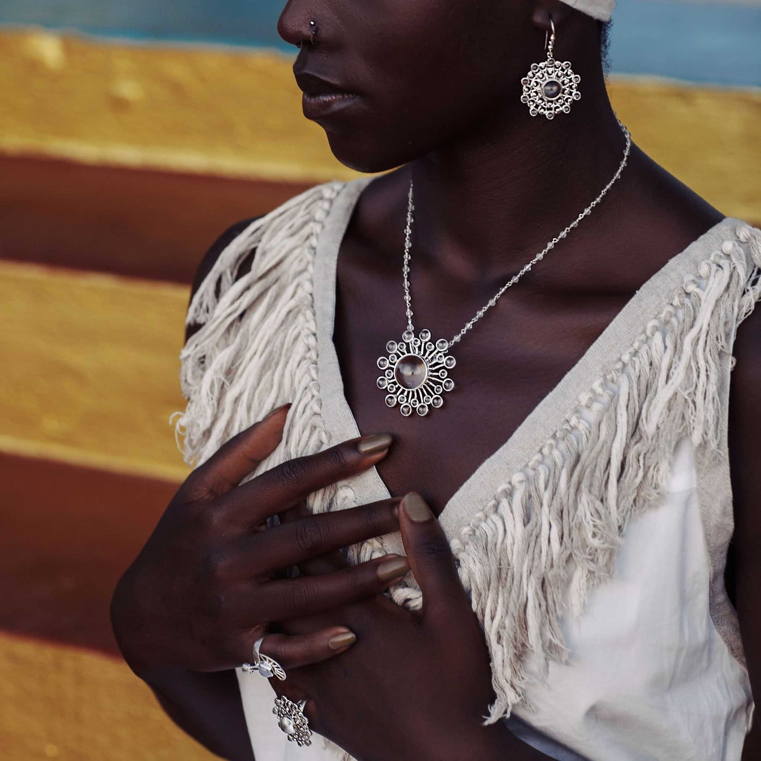 A person dressed in a white textured outfit that symbolizes purity is adorned with exquisite jewelry, including a striking pair of Purity Earhooks in sterling silver with crystal from Kaora Sandara Jewelry. The setting is enhanced by vibrant horizontal stripes in gradient hues of blue, yellow, and brown.