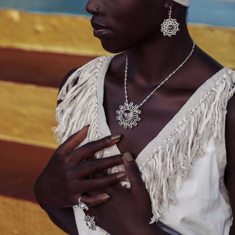 A person dressed in a white textured outfit that symbolizes purity is adorned with exquisite jewelry, including a striking pair of Purity Earhooks in sterling silver with crystal from Kaora Sandara Jewelry. The setting is enhanced by vibrant horizontal stripes in gradient hues of blue, yellow, and brown.