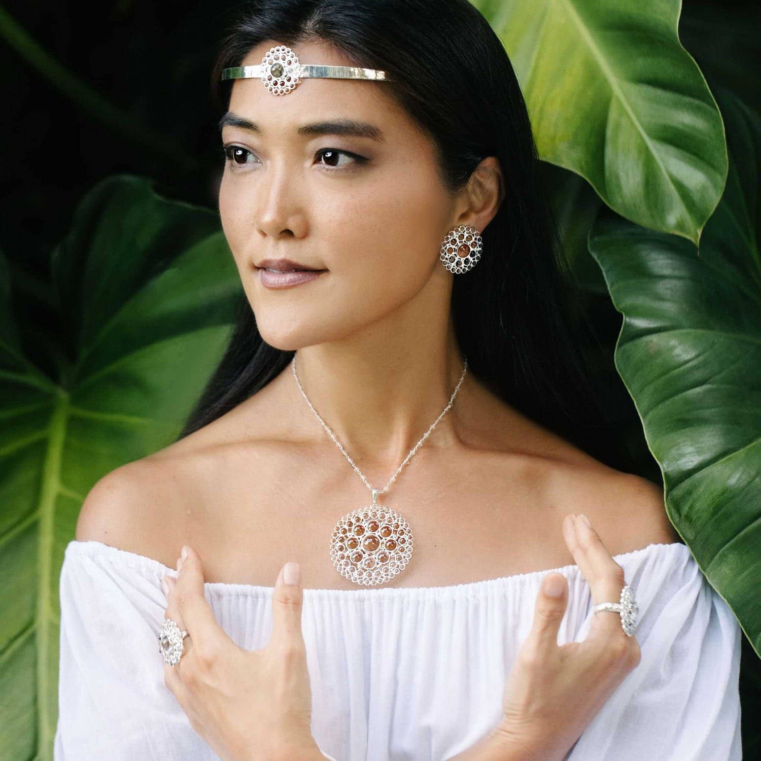 A woman adorned in Kaora Sandara Jewelry's Purity Pendant collection poses gracefully among lush green leaves, her intricate jewelry featuring transparent rhinestone crystals. She wears a sterling silver 925 necklace, earrings, ring, and headpiece. Her off-shoulder white top complements her long dark hair beautifully.