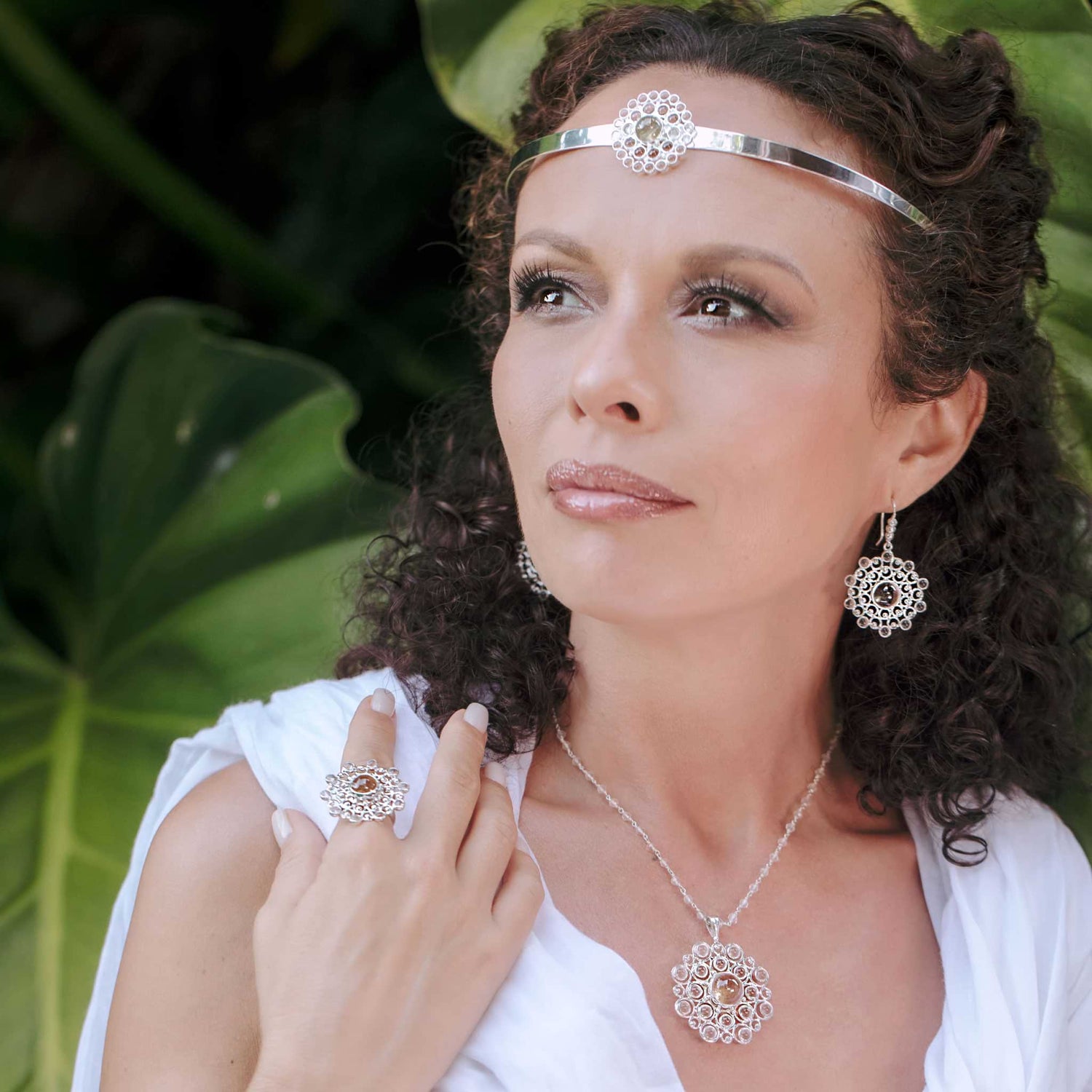 A woman with curly hair gazes into the distance, adorned in Kaora Sandara Jewelry's Sterling Silver 925 collection, featuring the Purity Headband enhanced with crystal, along with matching earrings, a necklace, and a ring. She stands gracefully against a backdrop of large green leaves.