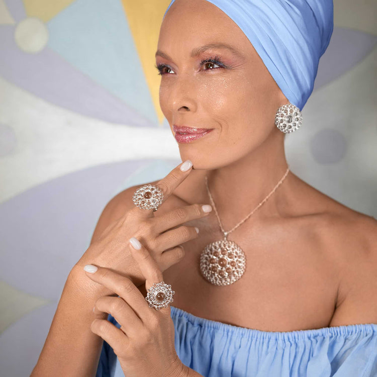 A woman wearing a blue headscarf and an off-the-shoulder top smiles subtly, elegantly showcasing Kaora Sandara Jewelry's Purity Earclips. These large, intricate pieces feature sterling silver with crystal spheres. The softly blurred background is adorned with pastel colors.