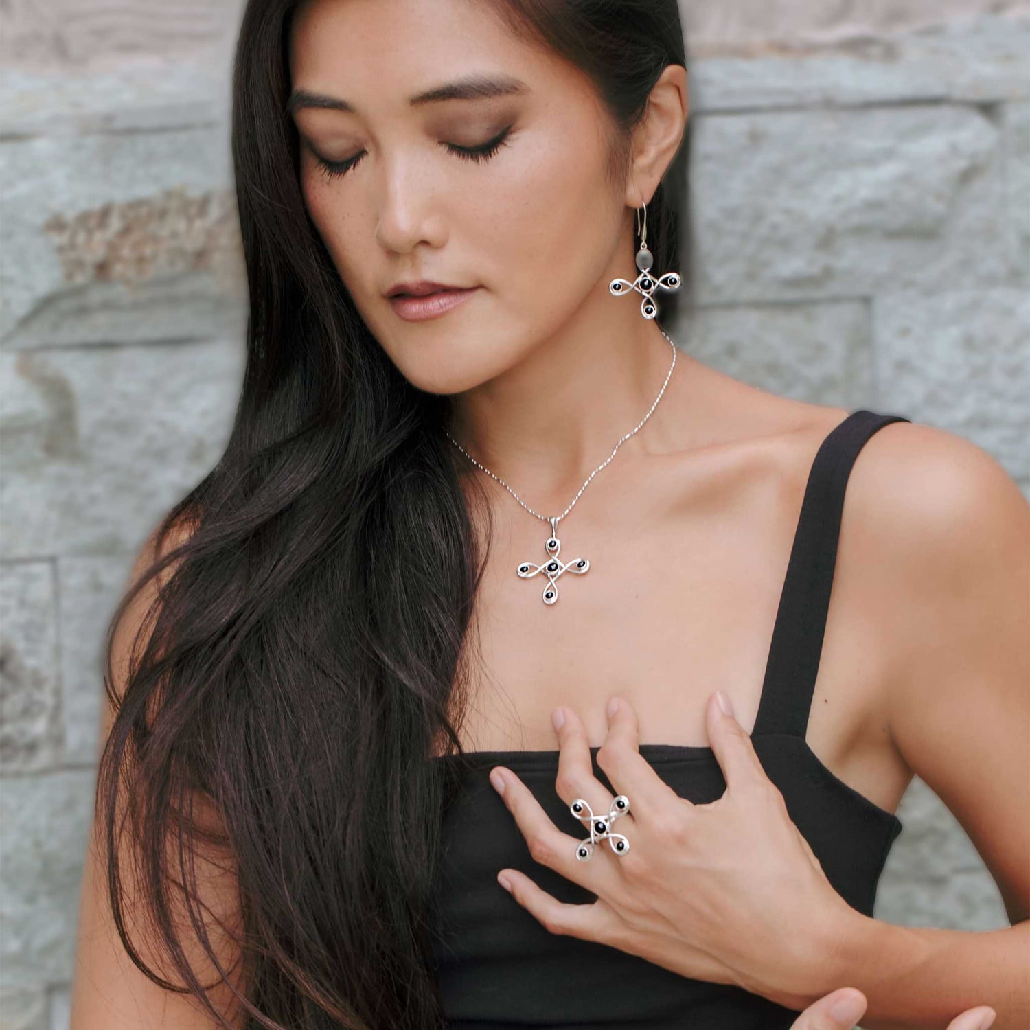 A woman with long, dark hair, wearing a sleek black sleeveless top, poses serenely with her eyes closed against a textured stone wall. She is adorned with Kaora Sandara Jewelry's Programs Clearing Pendant in sterling silver 925 featuring black agate stones, along with matching earrings and a ring.