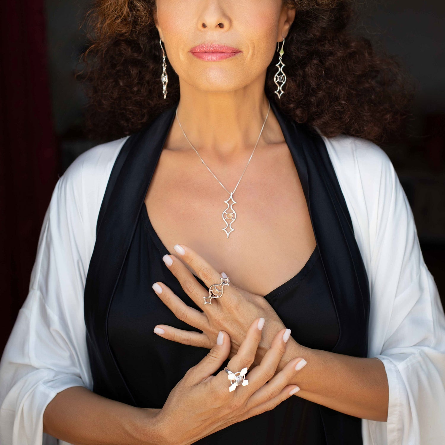 A person with curly hair elegantly displays Kaora Sandara Jewelry, wearing a stunning collection that features the Power's Activation Ring crafted from Sterling Silver with Crystal. Their hands gracefully rest over their chest, showcasing the jewelry. They are stylishly dressed in a black top complemented by a white shawl.