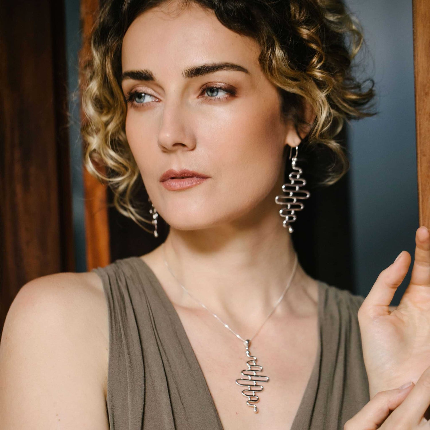 A woman with curly hair, wearing a sleeveless top and the elegant "Silver Earhooks Team Work" earrings by Kaora Sandara Jewelry, along with a matching necklace, gazes thoughtfully to the side. She stands near a wooden structure, softly lit to enhance her expression.