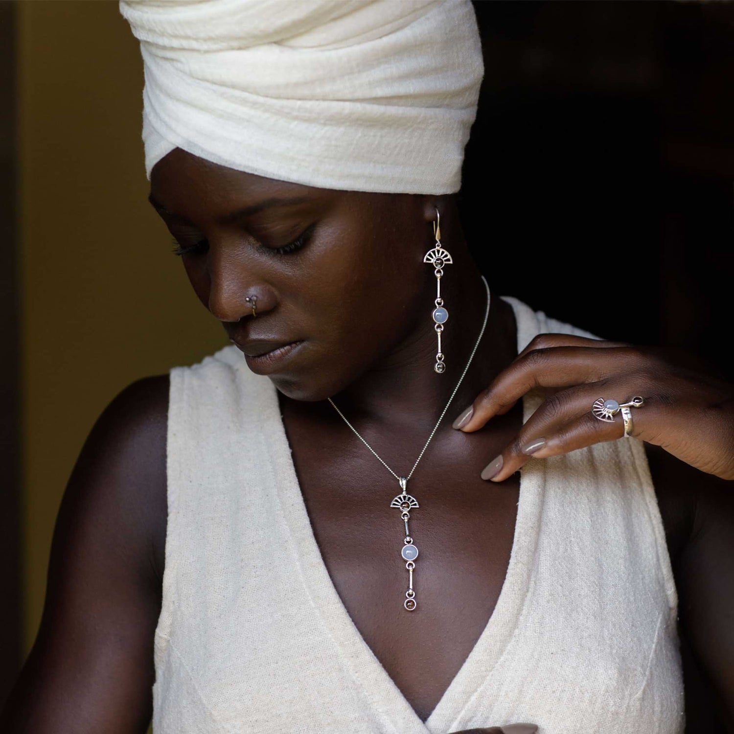 A person adorned in a white headwrap and sleeveless top elegantly displays intricate jewelry, including a long necklace, a dangling earhook, and a ring—all crafted by Kaora Sandara Jewelry in sterling silver with chalcedony. Their serene expression radiates peace of mind, embodying elegance and contemplation.