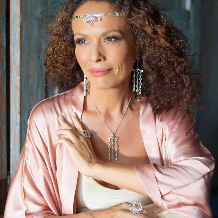 A woman with curly hair is elegantly dressed in a pink satin robe with silver jewelry, including a tiara, earrings, necklace, and rings. She sits indoors with a gentle smile, adorned by the Kaora Sandara Jewelry Magic Path Headband - Sterling Silver with Crystal. A rustic wooden door serves as the backdrop.