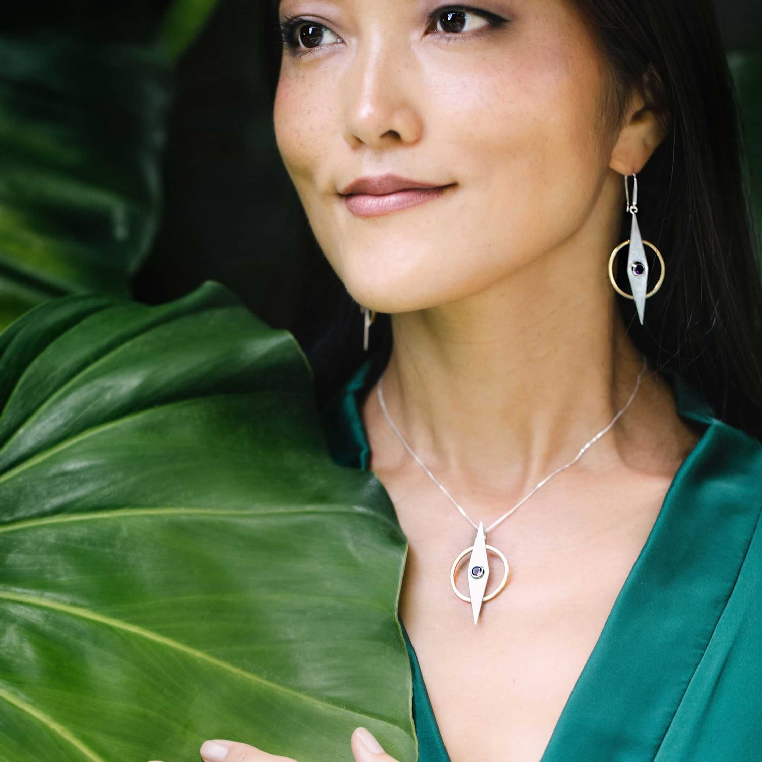 A woman in a green outfit is smiling softly, standing against large green leaves. She wears matching Kaora Sandara Jewelry, including Sterling Silver 925 earrings and an Intuition Pendant in Sterling Silver & Gold-Plated with Amethyst, featuring a circular and pointed design. The lush plant foliage creates a vibrant backdrop for her serene presence.