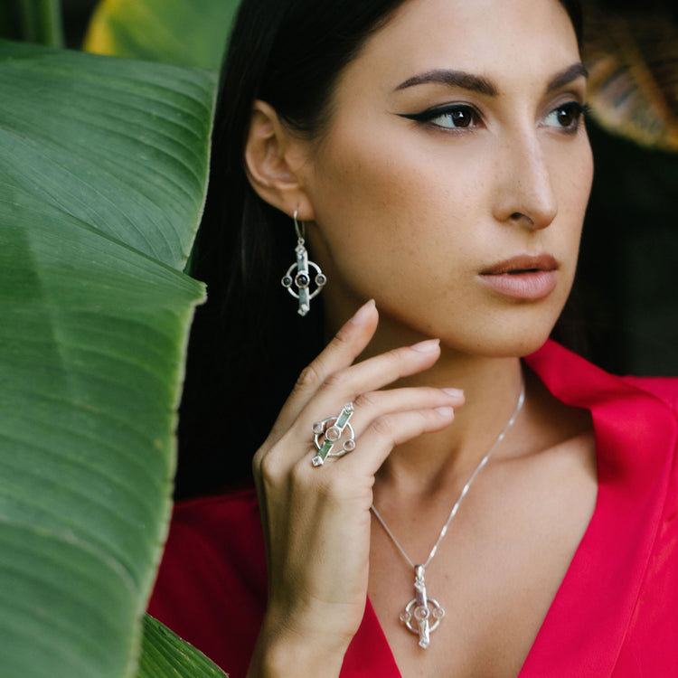 A woman in a red blouse poses against lush green leaves, embodying the essence of inner clarity. She is adorned with striking silver jewelry from Kaora Sandara Jewelry, featuring rhinestone crystal earrings, a ring, and the Inner Clarity Pendant made of sterling silver with crystal gemstones. Her dark and sleek hair is skillfully accentuated by bold eyeliner.