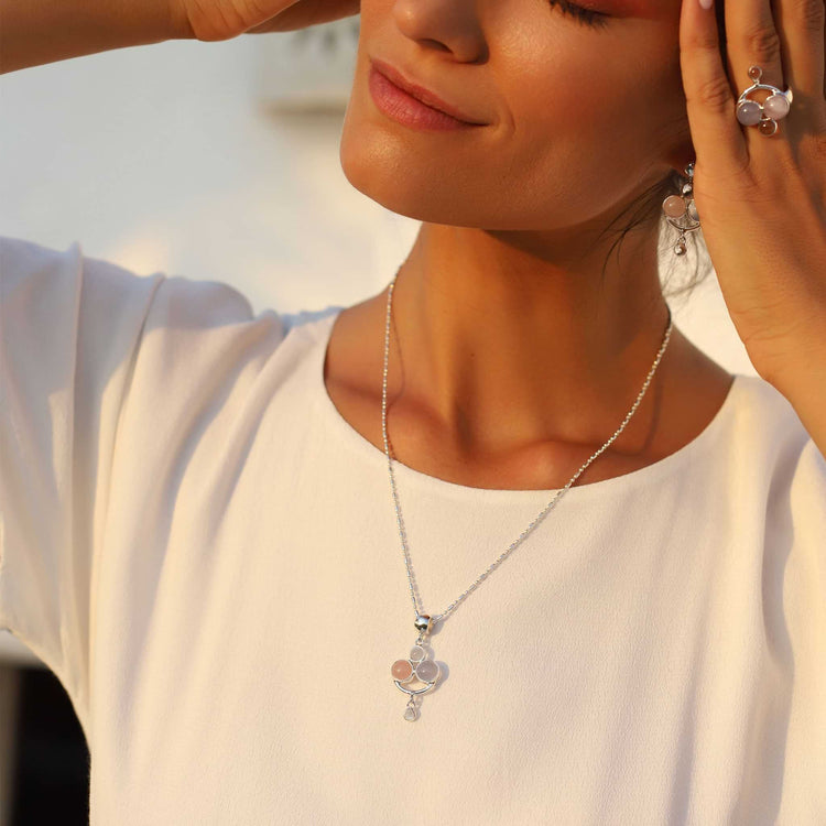 A woman in a white blouse gently touches her temple and cheek, showcasing elegant sterling silver jewelry by Kaora Sandara Jewelry. She wears the Happy Parents Pendant, beautifully crafted with crystal, chalcedony, and rose quartz, along with matching earrings and a ring that features similar designs. All the jewelry glistens warmly under the light, embodying the elegance of motherhood.