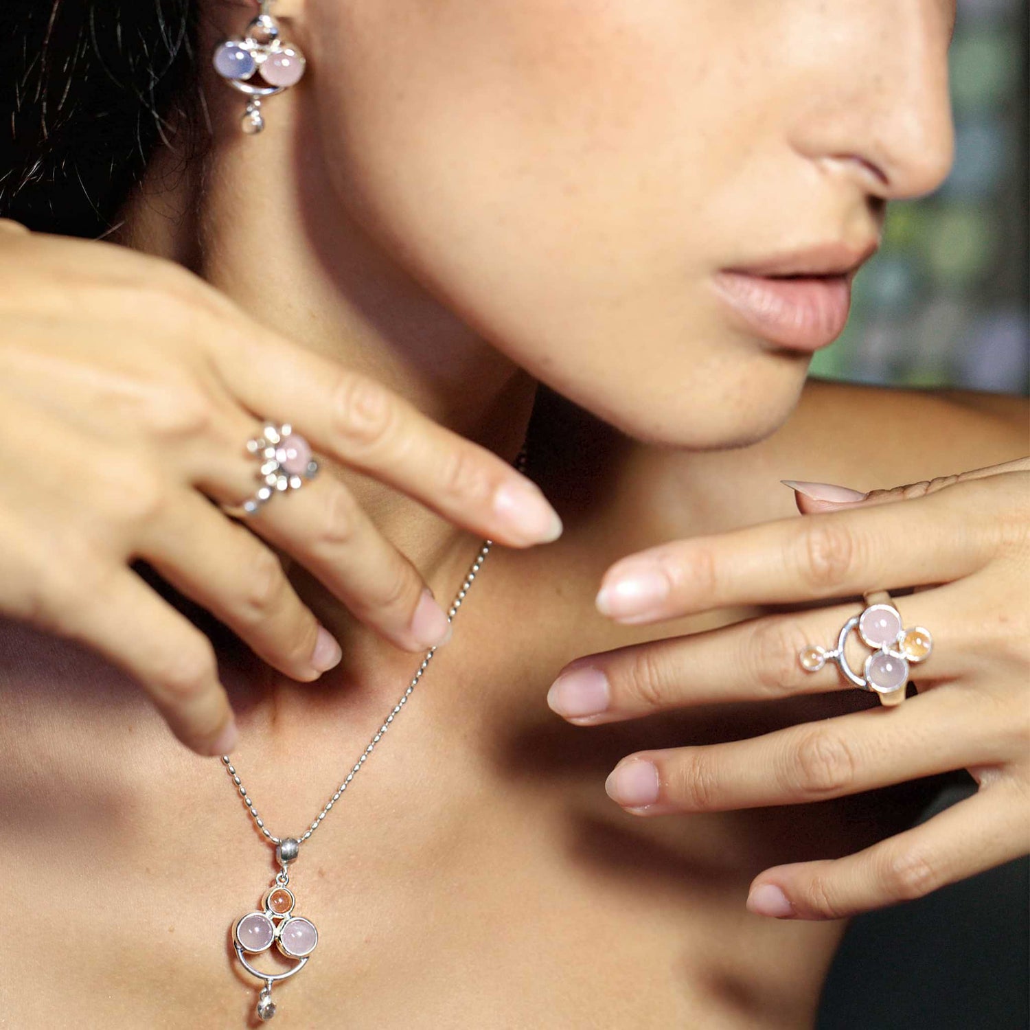 A woman wearing the Happy Parents Earstuds by Kaora Sandara Jewelry, crafted from sterling silver and adorned with light pink chalcedony and rose quartz, exudes a subtle healing effect. Her hands are elegantly positioned near her face, highlighting the earstuds against a blurred background.