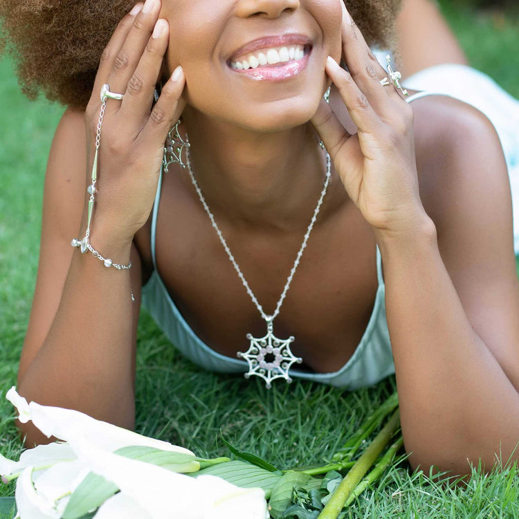 A woman lying on grass, smiling, adorns herself with Kaora Sandara Jewelry, showcasing the Freedom from the Ego Pendant in sterling silver. Her jewelry includes a star-shaped pendant featuring crystal and rose quartz and a matching bracelet. Her hands grace her face in a gesture of liberation, with rose quartz-colored lilies placed beside her.