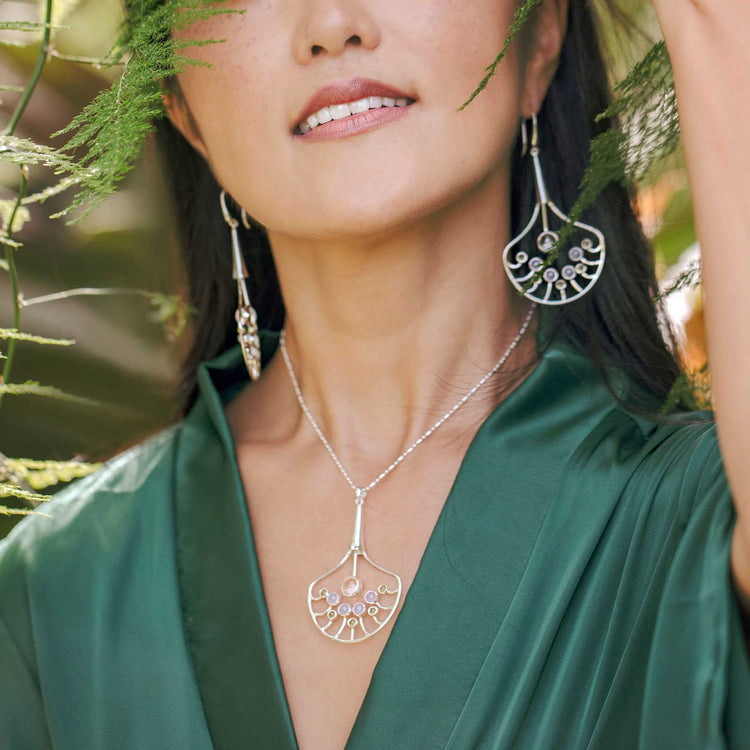 A woman wearing a green dress is partially obscured by leaves, showcasing silver jewelry from Kaora Sandara Jewelry. Her ensemble features the Body Relaxation Pendant in sterling silver, adorned with crystal, chalcedony, and peridot. Her gentle and serene expression complements the pendant's aura of energy flow and body relaxation.