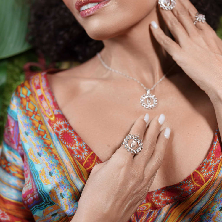 A woman with curly hair wears a colorful patterned dress. She is adorned with matching Kaora Sandara Jewelry in sterling silver 925, including a necklace, earrings, and the Anima Ring featuring circular designs. Her fingers gently touch her neckline and shoulder, where the Anima Ring - Sterling Silver with Crystal & Rose Quartz catches the light.