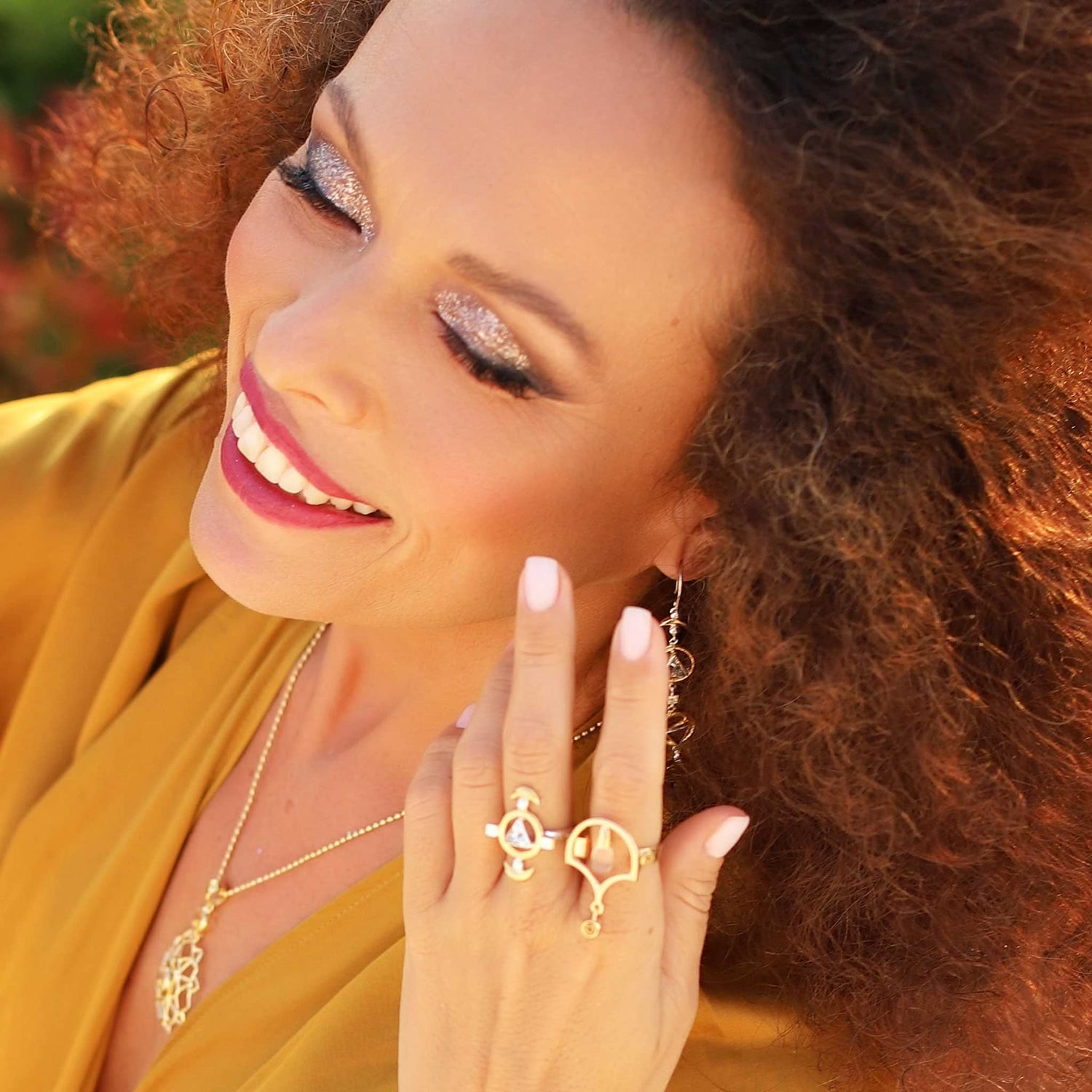 A woman with curly hair beams, adorned in gold jewelry and a stunning Citrine Sterling Silver necklace. Her sparkly eye makeup complements her yellow top, while a Kaora Sandara Jewelry Abundance Flow Ring – Sterling Silver & Gold-Plated with Crystal & Citrine glimmers on her finger. She exudes joy and relaxation.