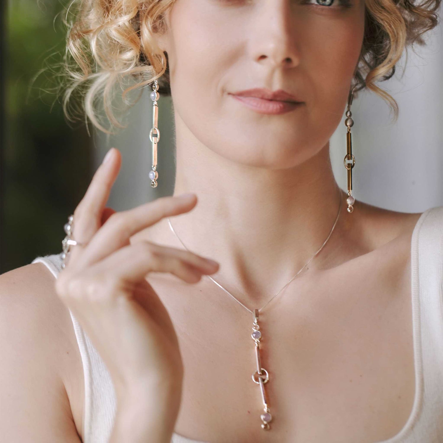 A person with curly hair, exuding tranquility, wears a white tank top paired with the State Pendant from Kaora Sandara Jewelry, crafted in sterling silver with crystal and chalcedony. They also sport long earrings featuring geometric designs while holding one hand close to their face against a blurred backdrop of greenery.