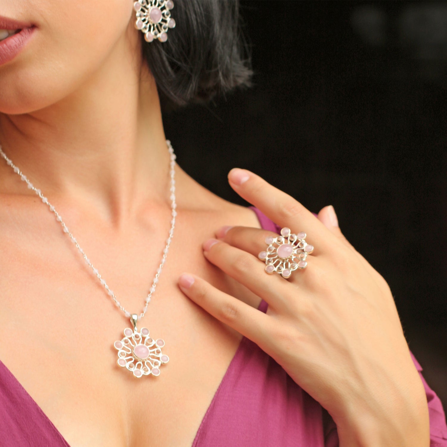 A woman wears a beautiful ensemble from Kaora Sandara Jewelry, featuring the Source of Love Ring in sterling silver adorned with crystals and rose quartz. Her outfit is complemented by matching earrings and a necklace showcasing intricate flower-like designs with small gemstones. Paired perfectly with her deep pink top, her hand gracefully hovers near the necklace.