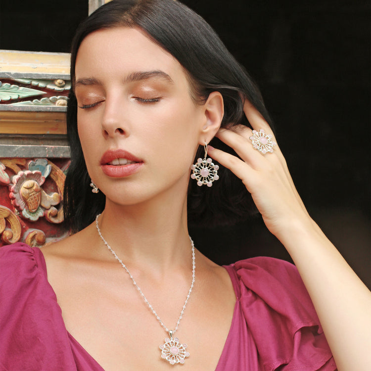 A woman with dark hair and closed eyes poses elegantly, adorned in a purple dress and intricate Kaora Sandara Jewelry. She is wearing the Silver Earhooks Source of Love, showcasing crystal and rose quartz gemstones, along with a ring and a necklace featuring a floral pattern against an ornate background.