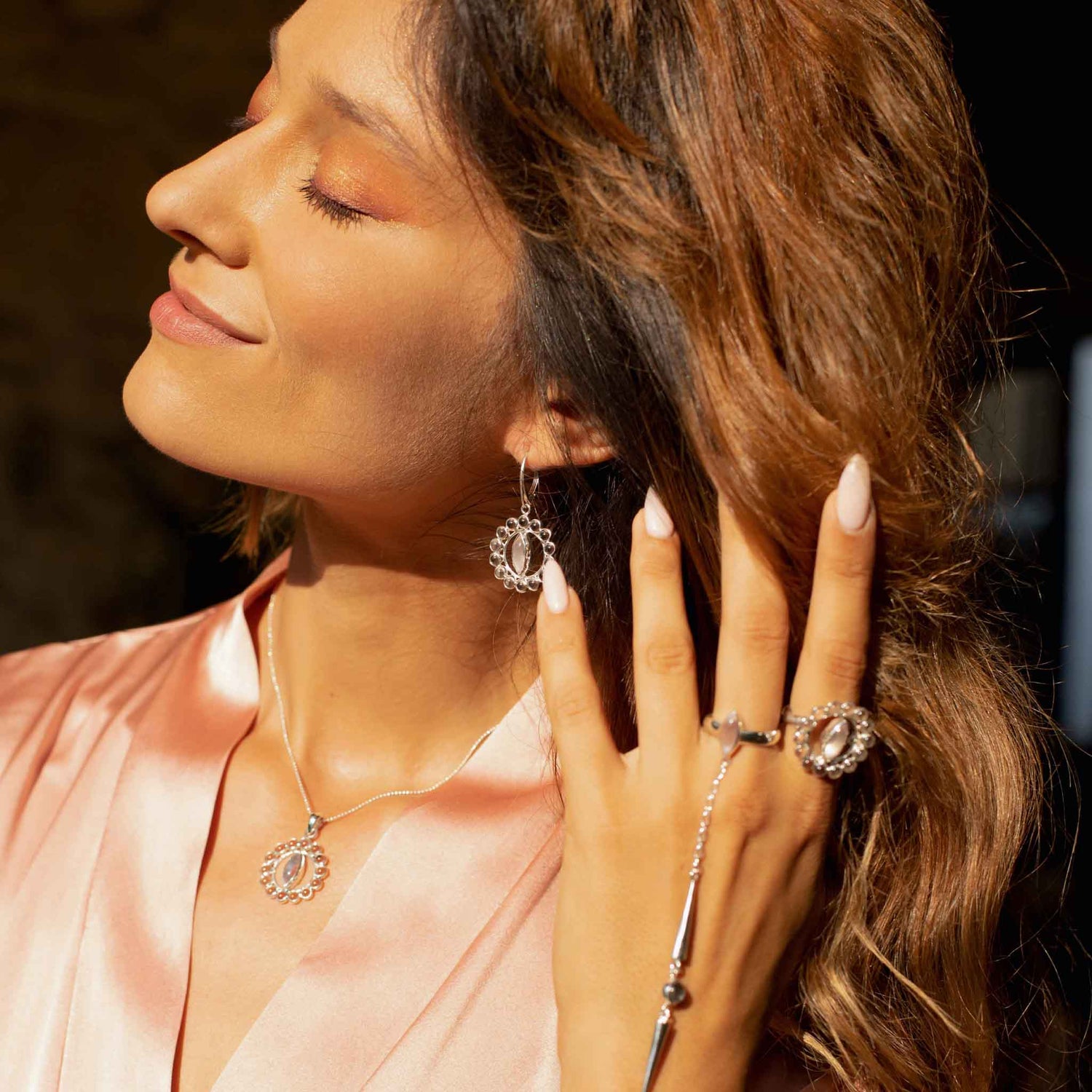 A woman with long wavy hair closes her eyes and smiles softly, showcasing elegant jewelry from Kaora Sandara Jewelry. She wears a pendant necklace, Anima Earhooks – Sterling Silver with Crystal & Rose Quartz dangling earrings, and a statement ring with intricate designs. Dressed in a silky pink top, she basks in warm light that catches the transparent rhinestone crystal accents.