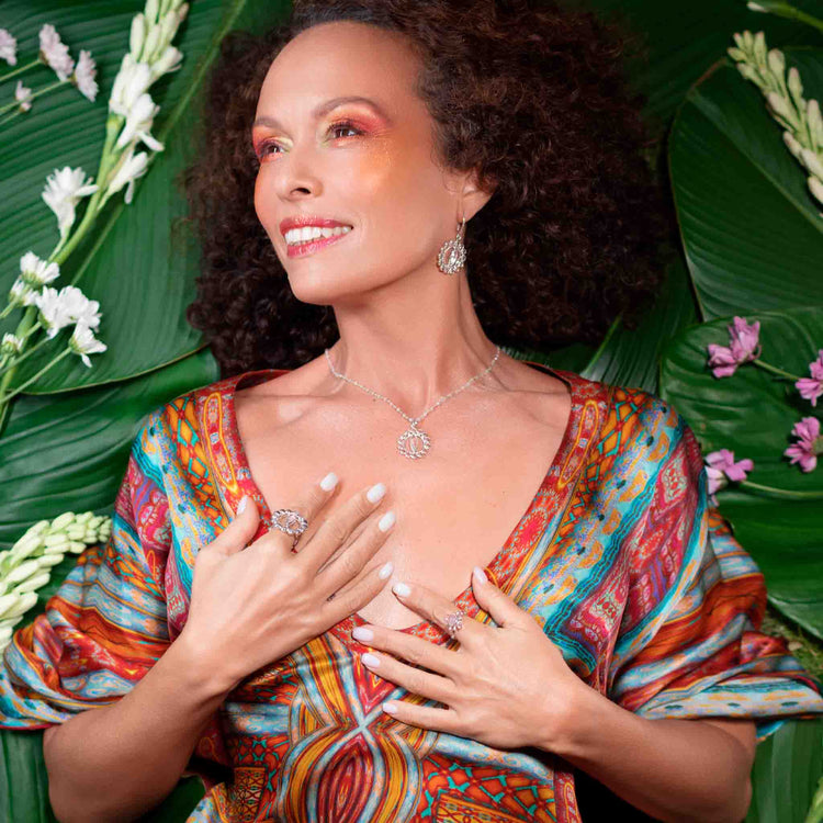 A woman with curly hair is lying on large green leaves, surrounded by white and pink flowers. She is wearing a brightly patterned dress, elegant earrings, rings, and the Anima Pendant from Kaora Sandara Jewelry – a sterling silver necklace featuring crystal and rose quartz. Her eyes are closed, and her hands rest peacefully on her chest with a serene expression.