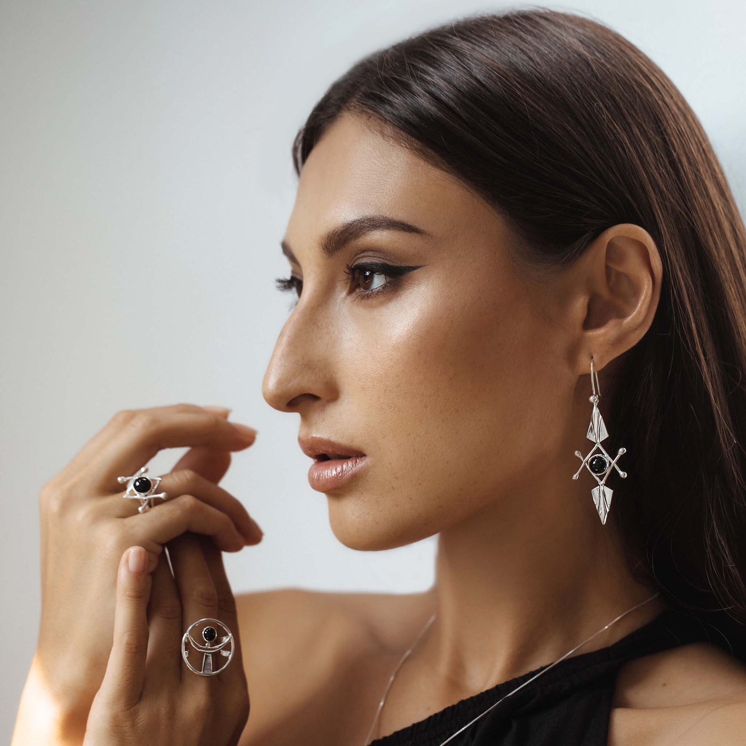 A woman with long dark hair wears intricate Kaora Sandara Jewelry, including Energy Protection Earhooks – Sterling Silver with Black Agate, a black agate ring, and a geometric pendant. She poses in profile against a light background, with her hands by her face, highlighting the elegant accessories designed for energy protection.