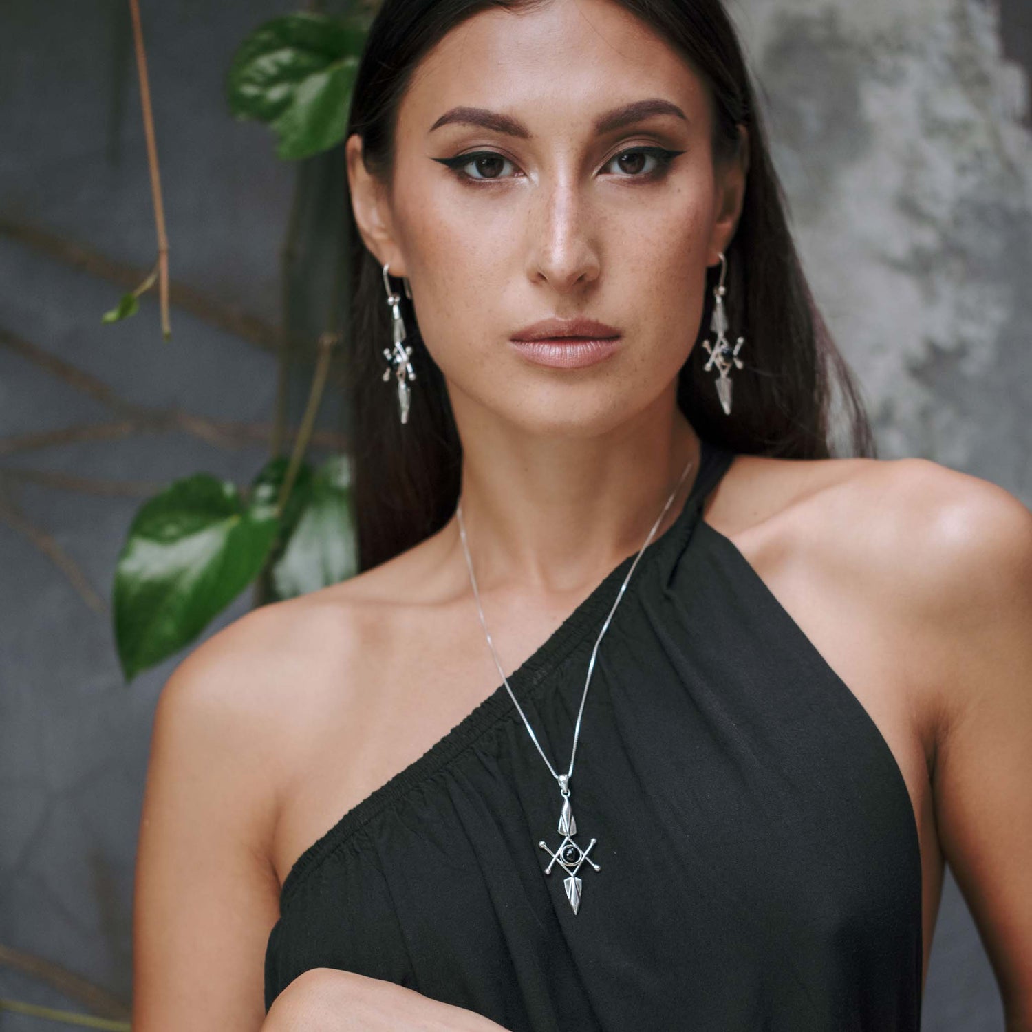 A woman with long dark hair wears a black one-shoulder dress and matching Sterling Silver 925 jewelry, including dangling earrings and a Kaora Sandara Jewelry Energy Protection Pendant - Sterling Silver with Black Agate, featuring intricate designs. She stands in front of a muted background with green leaves visible. A serious expression graces her face.