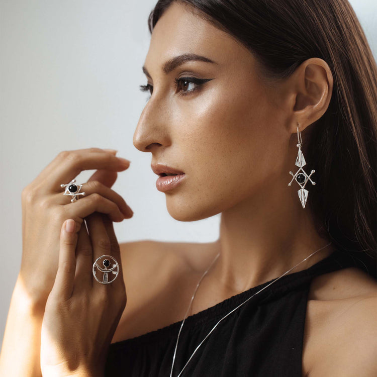 A woman with long brown hair is gazing to the left. She is wearing a black, off-shoulder top, long geometric drop earrings, and the Kaora Sandara Jewelry Energy Protection Ring - Sterling Silver with Black Agate alongside other rings with intricate designs. Her makeup is natural with a focus on eye makeup, and the background is softly lit.