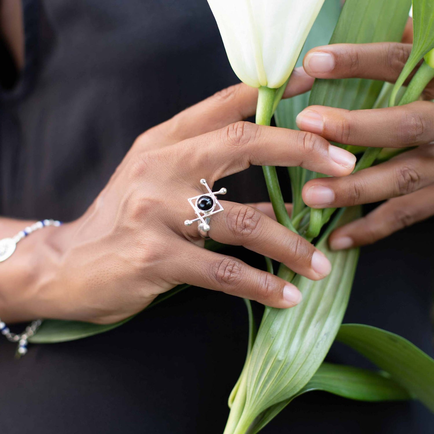 A person's hands hold a white lily. The left hand wears the Kaora Sandara Jewelry Energy Protection Ring, crafted from sterling silver 925 and featuring a black agate centerpiece. Dressed in a black outfit, the person has a thin bracelet adorning their right wrist. The green leaves of the lily contrast beautifully with the jewelry and skin tones.