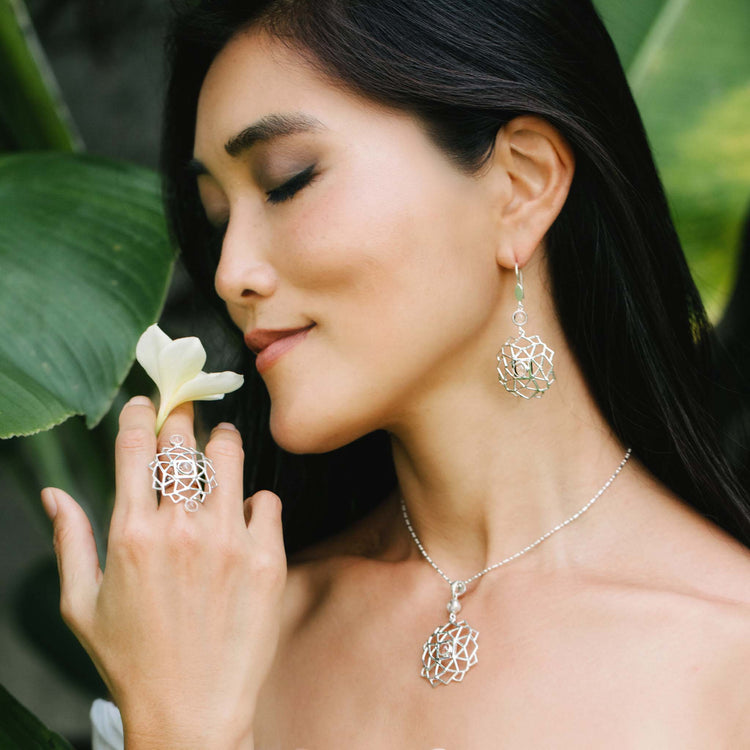 A woman with long dark hair smiles gently, her eyes closed, while wearing matching Kaora Sandara Jewelry: a set of sterling silver pieces including an intricate geometric necklace, earrings, and the stunning Expansion Ring adorned with crystal gemstones. She holds a white flower near her face, standing against a backdrop of large green leaves in an elegant transition of natural beauty.