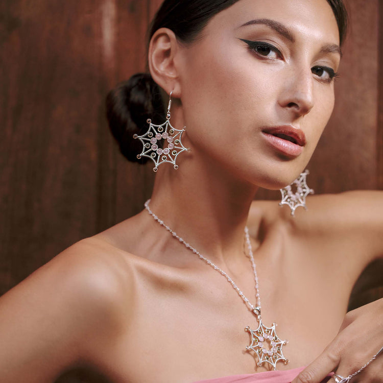 A woman with winged eyeliner and a sleek updo poses against a wooden background. She is wearing intricate web-like earrings, paired with the Freedom from the Ego Pendant by Kaora Sandara Jewelry, both adorned with delicate sterling silver 925 geometric designs. She is dressed in a strapless pink top.
