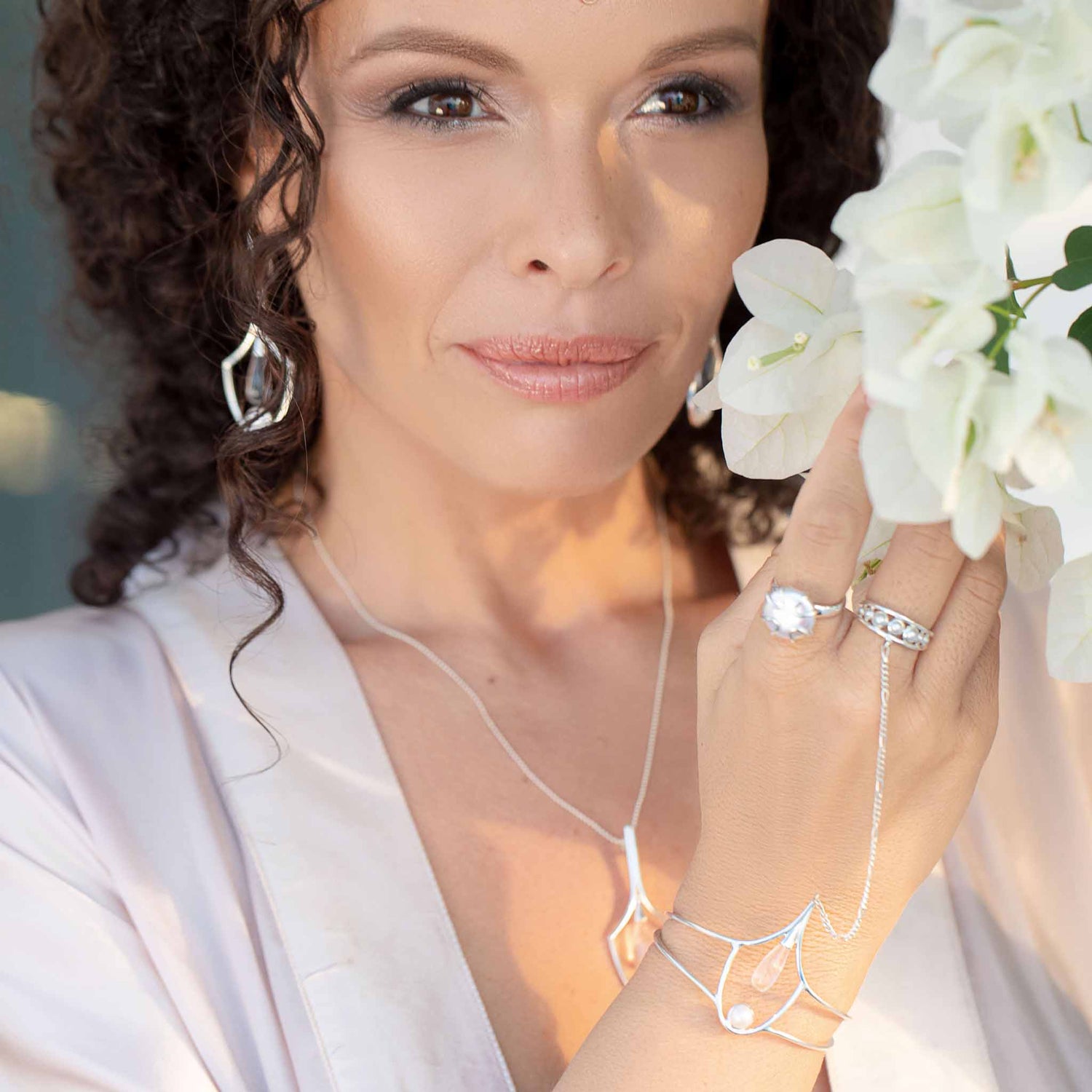 A woman with wavy hair is adorned with elegant silver jewelry from Kaora Sandara Jewelry, including rhinestone crystal earrings, rings, and the Silver Hand Chain Youth Key with Crystal & Pearl. She holds white flowers close to her face with a serene expression that highlights her youthful beauty. She is wearing a light-colored blouse.