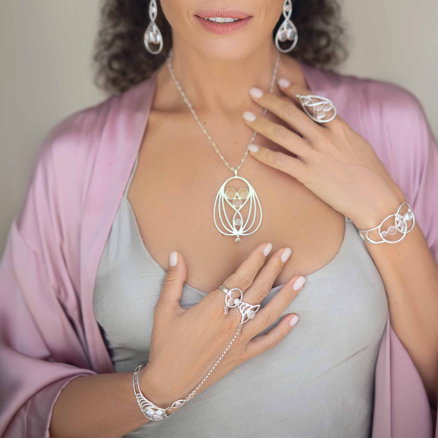 A person adorned with a stunning silver necklace featuring an intricate design, accompanied by matching silver earrings, and various Sterling Silver 925 rings and bracelets. The person is dressed in a light gray top and a light pink robe, with one hand graced by a Rose Quartz ring and other accessories from Kaora Sandara Jewelry, including the elegant Silver Hand Chain Transition Gift with Crystal & Rose Quartz Sphere Gemstones. One hand rests on their chest while the other is positioned near their waist.
