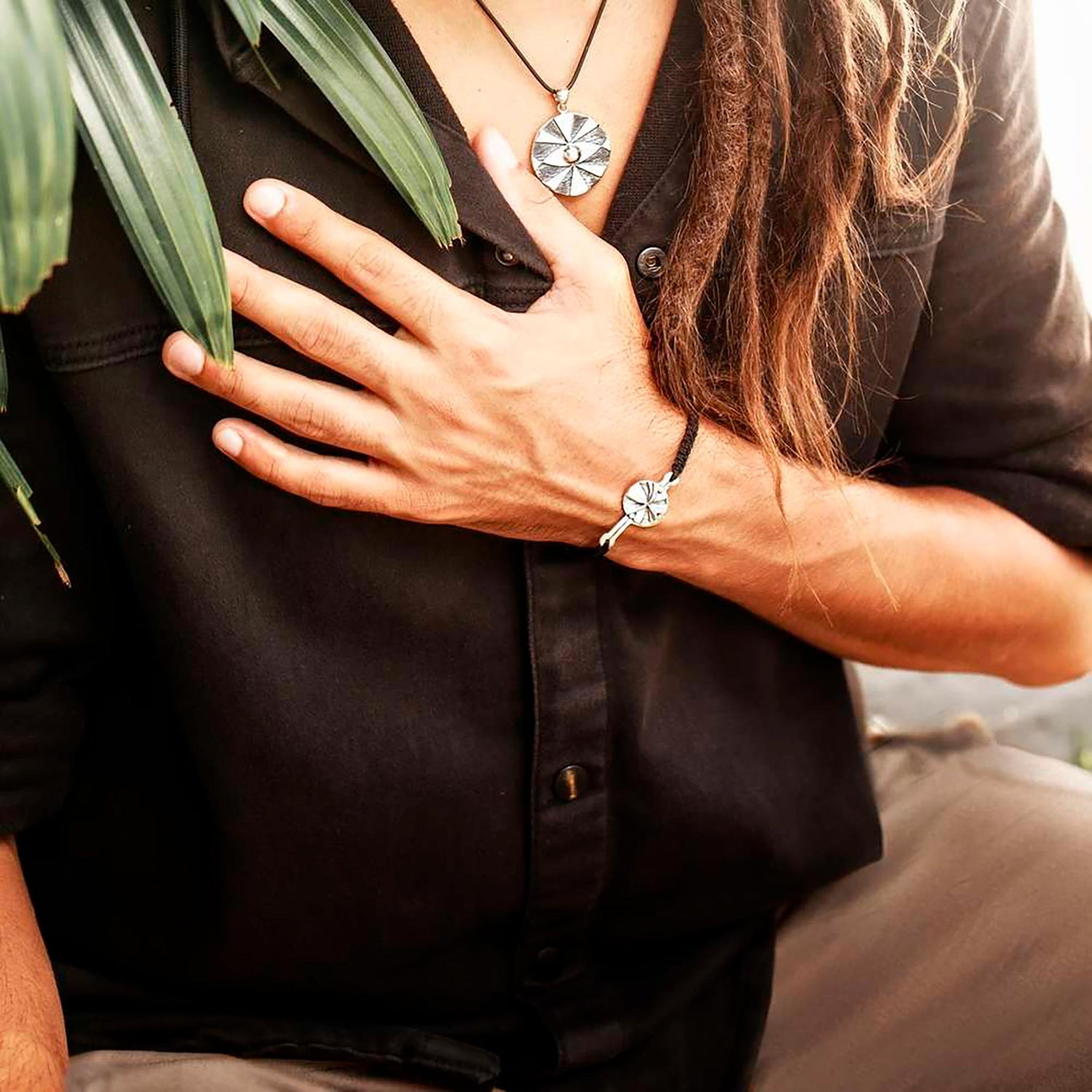 A person adorned in a black shirt, showcasing a necklace with a circular sterling silver 925 pendant, gently rests their hand on their chest. The ensemble includes the Kaora Sandara Jewelry Silver Male Bracelet on a Dark Grey String with the Code Abundance, which mirrors the necklace's design. A green leafy plant, representing financial prosperity, is partially draped over their shoulder.