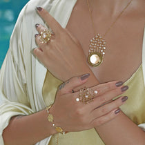 A person is adorned with a gold necklace and circular pendant with pearls, complemented by two studded rings from Kaora Sandara Jewelry's Ring Expansion collection. A matching gold bracelet completes the look against light, silky fabric.