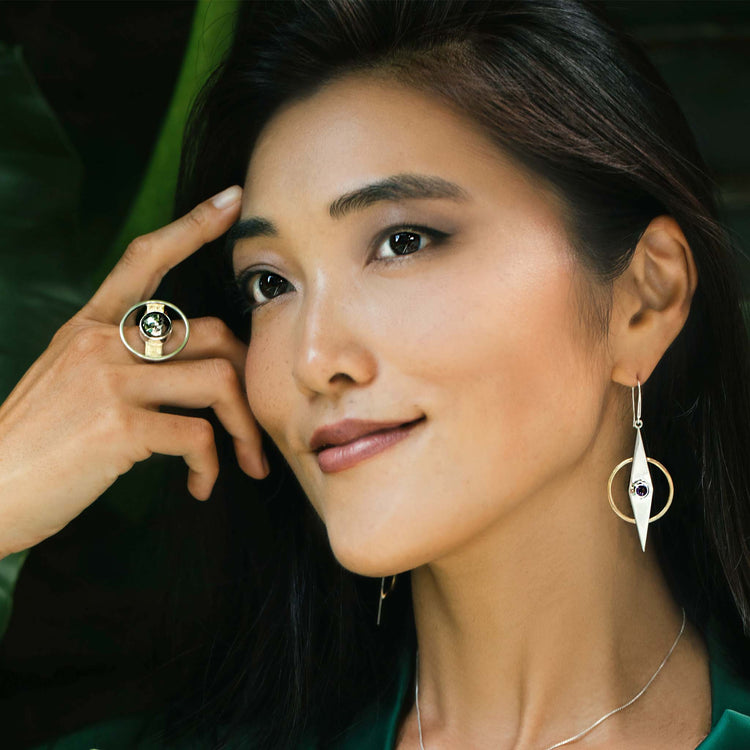 A woman with long dark hair smiles softly while touching her forehead with one hand. She wears a large Animus Ring – Sterling Silver & Gold-Plated with Pyrite from Kaora Sandara Jewelry and matching drop earrings featuring circular and elongated shapes, with a green background behind her.