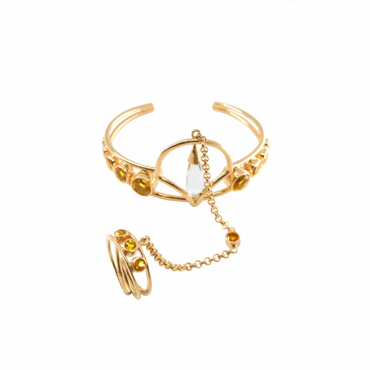 Against a white background, there is a hand ornament from the Abundance Flow series, made of silver and gold-plated. It consists of a bracelet and a ring connected by a chain. The ring features citrines, while the bracelet showcases a drop-shaped rock crystal and several citrines.