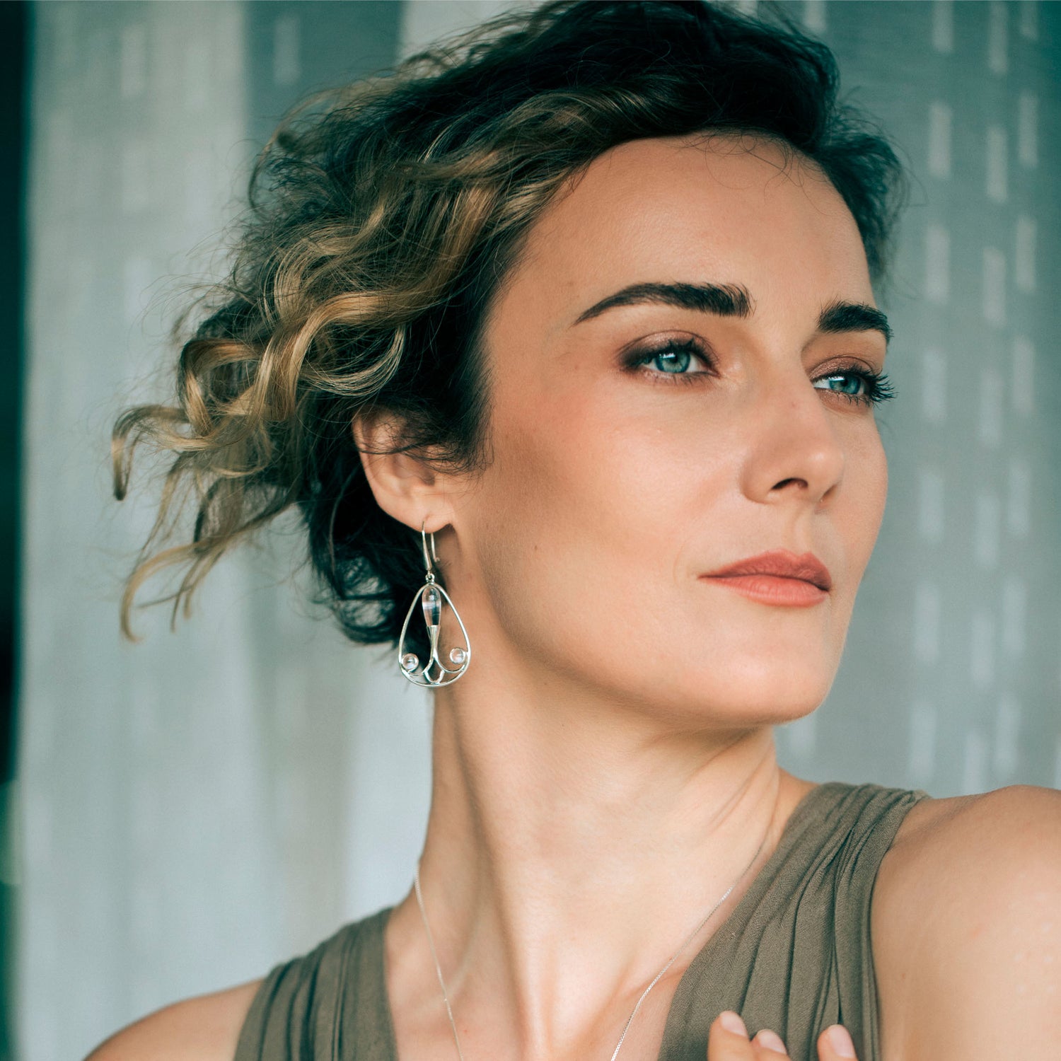 A person with short, wavy hair looks away from the camera with a neutral expression. They are wearing a beige sleeveless top and Silver Earhooks Self-Worth – Crystal Gemstone earrings by Kaora Sandara Jewelry that dangle gracefully, exuding individuality against the blurred background.