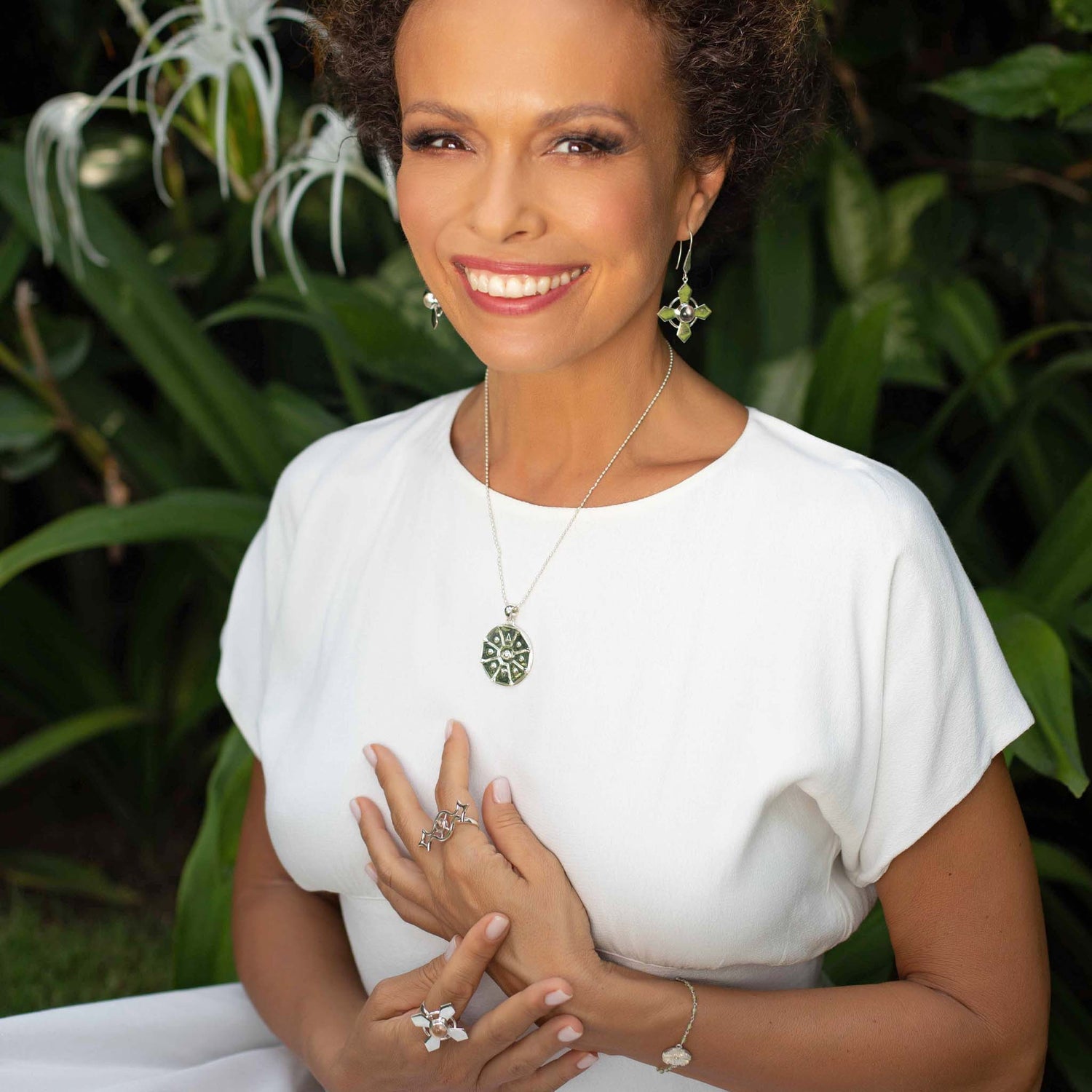 A woman wearing a white dress smiles at the camera. She is adorned with green jewelry from Kaora Sandara Jewelry, including Power's Activation Earhooks in sterling silver with transparent rhinestone crystals, as well as earrings, rings, and a bracelet. The background shows lush green plants with white flowers.