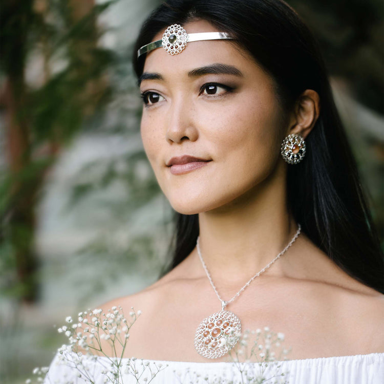 A woman in an outdoor setting is adorned with an elaborate headband featuring a circular design at the center, matching sterling silver 925 earrings, and the Kaora Sandara Jewelry Purity Pendant - Sterling Silver with Crystal in Size L. She is dressed in an off-the-shoulder white top, holding delicate white flowers, and gazing ahead. The background consists of blurred greenery.