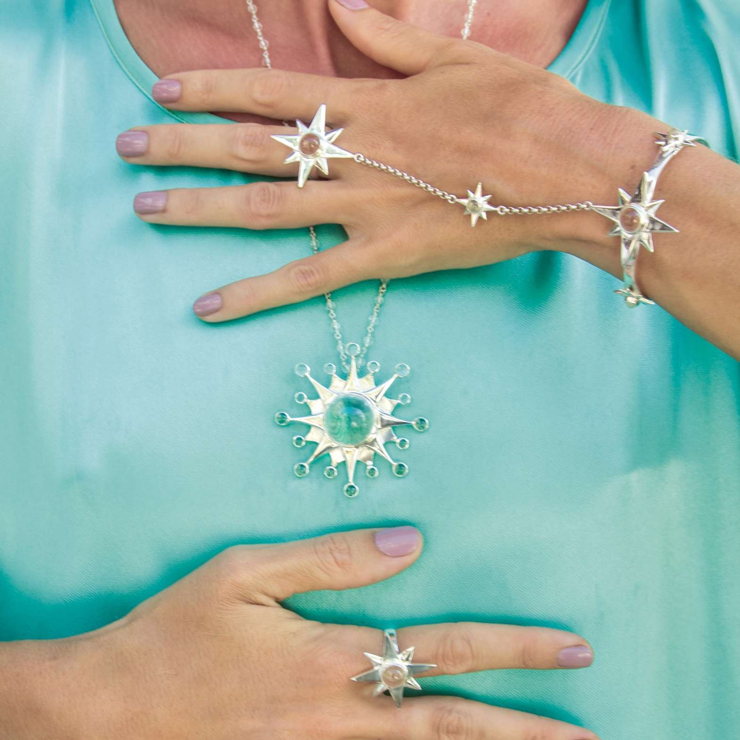 Close-up of a person wearing matching star-shaped jewelry from Kaora Sandara Jewelry, featuring the Silver Hand Chain Magic Star with Crystal Sphere Gemstones. The pieces, made of Rock crystal and Sterling silver 925, include a necklace, bracelet, ring, and distinctive hand jewelry with chains connecting the bracelet to the ring. The circular stone centers are designed to enhance intuition and sensory perception when worn over turquoise satin or silk garments.