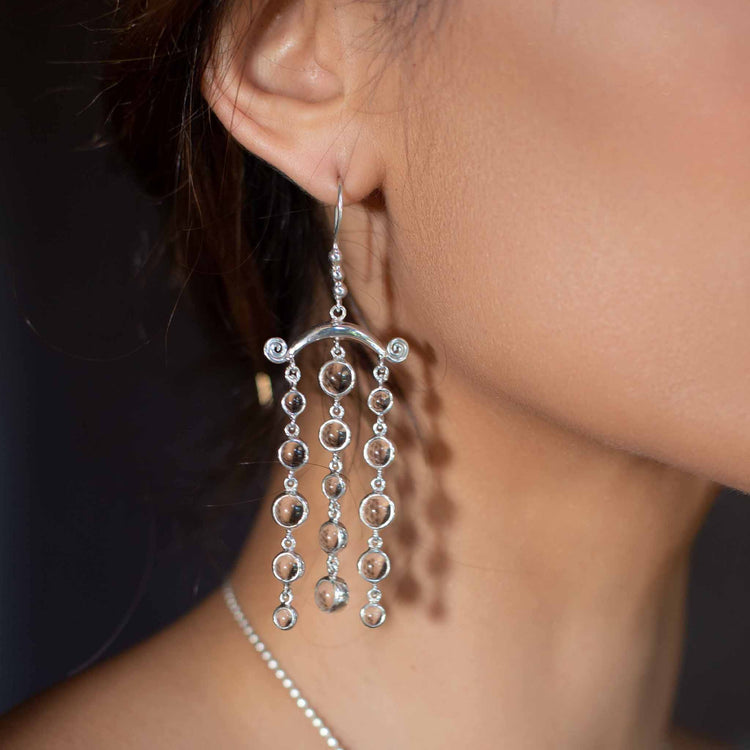 Close-up of a person wearing elegant Magic Path Earhooks – Sterling Silver with Crystal Gemstones by Kaora Sandara Jewelry. The earhooks feature multiple cascading chains of small circular links and transparent rhinestone crystals, hanging from a curved structure. The person has fair skin and their dark hair is tied back, partly visible.