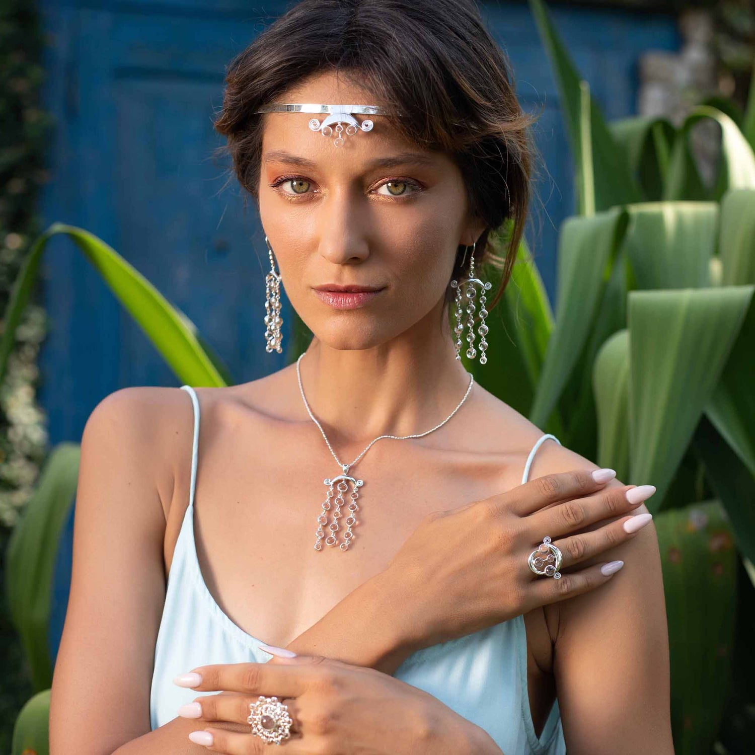 A woman with short brown hair is wearing a delicate headpiece, long dangling Magic Path Earhooks – Sterling Silver with Crystal Gemstones by Kaora Sandara Jewelry, a necklace, and multiple Sterling Silver 925 rings with intricate designs. She is dressed in a light blue sleeveless top and stands in front of green plants and a blue door.